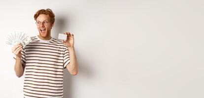 Shopping and finance concept. Cheerful handsome redhead man showing plastic credit card and cash, smiling amazed, white background photo