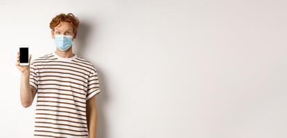 Covid-19, online doctor concept. Smiling redhead man in face mask showing smartphone screen, standing over white background photo
