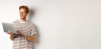 Handsome redhead man working freelance on laptop, smiling and looking pleased at computer screen, white background photo
