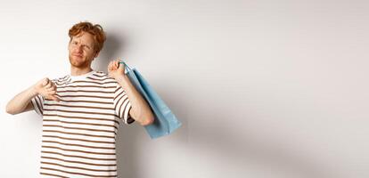 Disappointed young man with red hair and beard showing thumbs-down after bad shopping experience, holding bag over shoulder and frowning upset, white background photo
