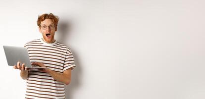 Impressed young man holding laptop in hands, staring at camera with excited face, reading online promotion on website, standing against white backround photo