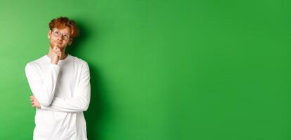 Thoughtful redhead man in glasses making choice, looking up and thinking, standing over green background photo
