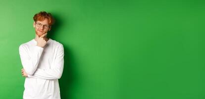 Intrigued young man with red hair, wearing glasses, looking at camera pleased and thoughtful, having an idea, standing over green background photo