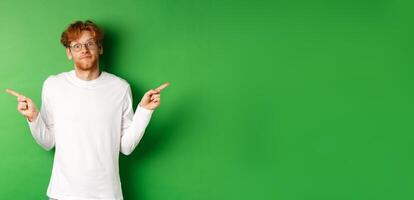 Indecisive funny guy with red hair pointing fingers sideways, staring confused at camera and showing two promo offers, standing over green background photo