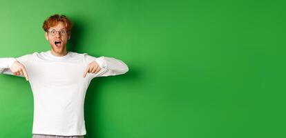 Surprised and excited young redhead man scream of joy and amazement, checking out awesome promo, pointing fingers down, standing over green background photo