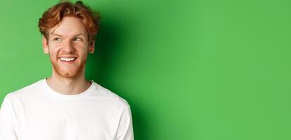 Headshot of happy redhead man with beard wearing white long sleeve, looking left at copy space and smiling, standing over green background photo