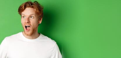 Close up of surprised and impressed redhead man checking out promotion offer, looking left with dropped jaw, standing over green background photo