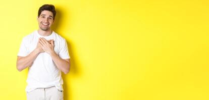 Image of grateful handsome guy in white t-shirt, holding hands on heart and smiling pleased, express gratitude, thanking for something, standing over yellow background photo