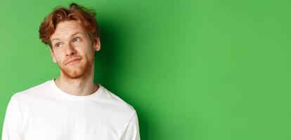 Smiling redhead man with messy haircut and beard tilt head, looking left with pleased face, standing over green background photo