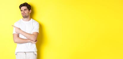 Unimpressed young man complaining, pointing and looking with skeptical face at bad product, standing against yellow background photo