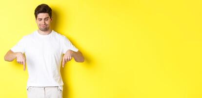 Skeptical young man in white t-shirt, pointing and looking down upset, disapprove and dislike product, standing over yellow background photo