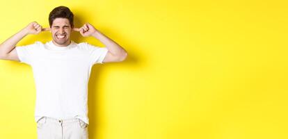 Annoyed man grimacing and shutting ears, complaining on loud noise, standing against yellow background photo