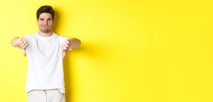 Skeptical guy showing thumbs down and smirking, dislike and disagree, standing over yellow background photo