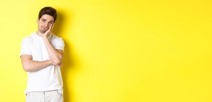 Bored and unamused man looking with indifference, standing in white clothes over yellow background photo