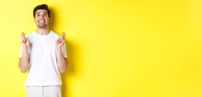 Nervous and hopeful man praying to god, making wish with fingers crossed, panicking and standing over yellow background photo