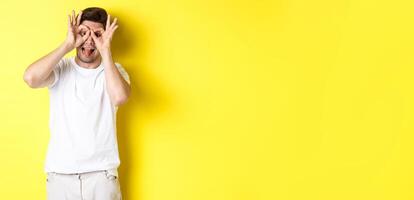 Young man showing funny faces and sticking tongue, standing playful against yellow background photo