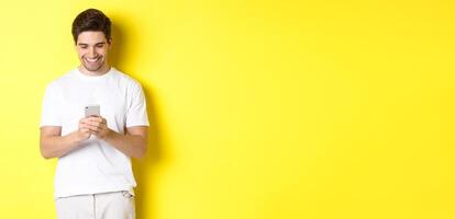 Young man reading text message on smartphone, looking at mobile phone screen and smiling, standing in white t-shirt against yellow background photo
