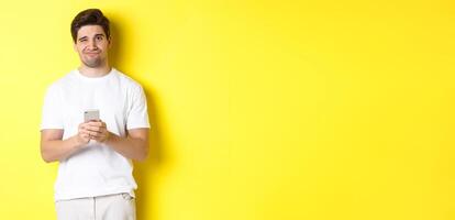 Displeased and reluctant man grimacing, being unamused by message on smartphone, standing over yellow background photo