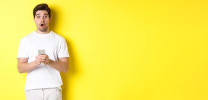 Man looking surprised, using smartphone, open mouth and saying wow, standing against yellow background photo
