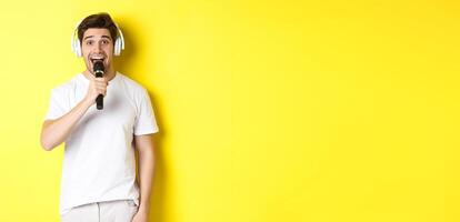 Man in headphones holding microphone, singing karaoke song, standing over yellow background in white clothes photo