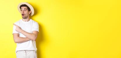 Concept of tourism and lifestyle. Excited handsome guy in straw hat checking out advertisement, pointing and looking at upper left corner logo, yellow background photo