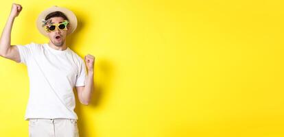Concept of tourism and lifestyle. Happy guy tourist enjoying trip, rooting for you, fist pump and triumphing, going on journey in summer hat and sunglasses, yellow background photo