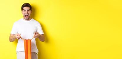 Surprised guy open shopping bag with fist, looking excited and happy at camera, standing against yellow background photo