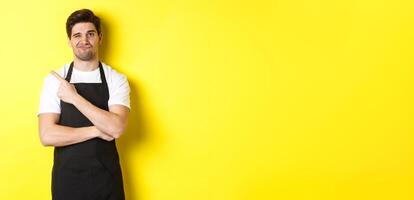 Displeased barista in black apron pointing finger at upper left corner, grimacing skeptical, dislike something, standing against yellow background photo