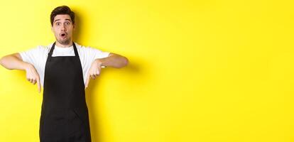 Surprised seller in black apron pointing fingers down, looking amazed at camera, standing over yellow background photo