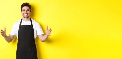 Smiling waiter in black apron holding your logo or box, spread hands as if carry something large, standing over yellow background photo