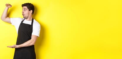 Waiter in black apron looking amazed at something large, holding big object, standing over yellow background photo