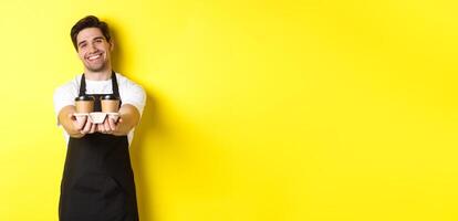 Friendly barista in black apron giving takeaway order, holding two cups of coffee and smiling, standing over yellow background photo
