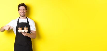 Handsome barista holding two cups of takeaway coffee, pointing finger at drinks and smiling, standing in black apron against yellow background photo