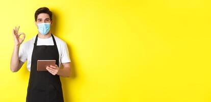 Concept of covid-19, small business and pandemic. Salesman in medical mask and black apron showing ok sign, taking orders with digital tablet, yellow background photo