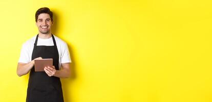 Handsome waiter taking orders, holding digital tablet and smiling, wearing black apron uniform, standing over yellow background photo
