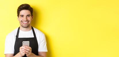 Close-up of handsome barista sending message on mobile phone, smiling happy, standing over yellow background photo