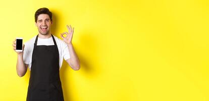 Handsome coffee shop worker showing ok sign and smartphone screen, recommending application, standing over yellow background photo