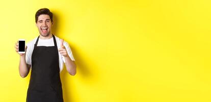 Happy waiter showing mobile screen and thumb up, recommending cafe restaurant app, standing over yellow background photo