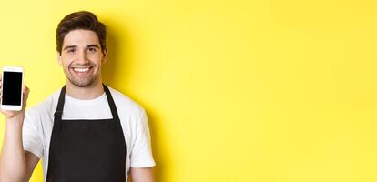 Close-up of handsome waiter in black apron showing smartphone screen, recommending app, standing over yellow background photo