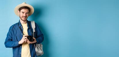 Handsome guy going on summer vacation, backpacking on holiday. Tourist with straw hat and camera smiling happy, standing on blue background photo