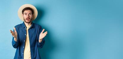 Shocked male tourist on vacation raising hands up and looking at camera startled, react to big news with disbelief, standing on blue background in straw summer hat and shirt photo