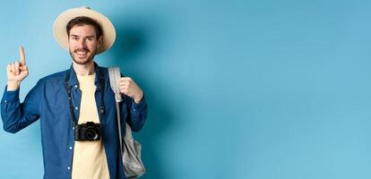Handsome happy tourist in summer hat, holding backpack and camera, pointing finger up at logo, recommending travel agency or place on vacation, blue background photo