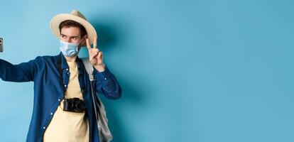 Covid-19, pandemic and travel concept. Happy and positive guy in tourist hat taking selfie and showing peace sign, posing near sightseeing, blue background photo