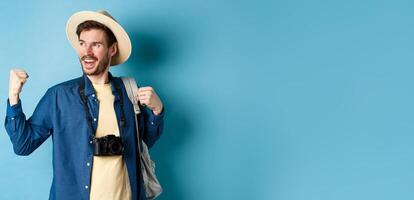 Cheerful tourist rejoicing on vacation, celebrating summer holidays, showing fist pump gesture and saying yes with satisfied face, looking aside, blue background photo