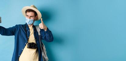 Covid-19, pandemic and travel concept. Happy male tourist taking pictures on vacation, make selfie with thumb up, recommending hotel, standing on blue background photo