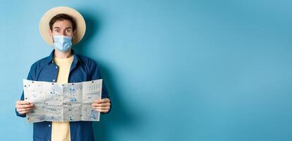 Covid-19, pandemic and travel concept. Tourist in medical mask look surprised, holding map, standing on blue background photo
