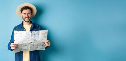 Happy and positive guy on vacation, looking at camera and holding map, smiling excited, going on summer travel, standing on blue background photo