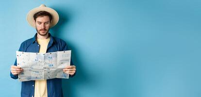 Handsome tourist with summer hat reading map, looking for sightseeing on vacation, standing on blue background photo