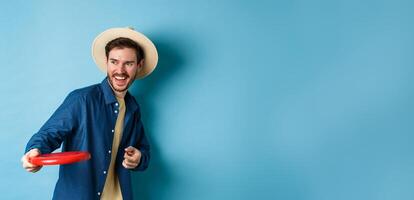 Happy man laughing and playing frisbee, throwing at friend and smiling, standing in summer hat on blue background photo