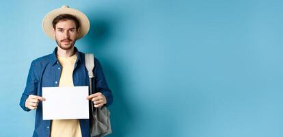 Image of smiling guy in straw hat backpacking, hitchhiking with piece of paper, travelling abroad on summer vacation, blue background photo